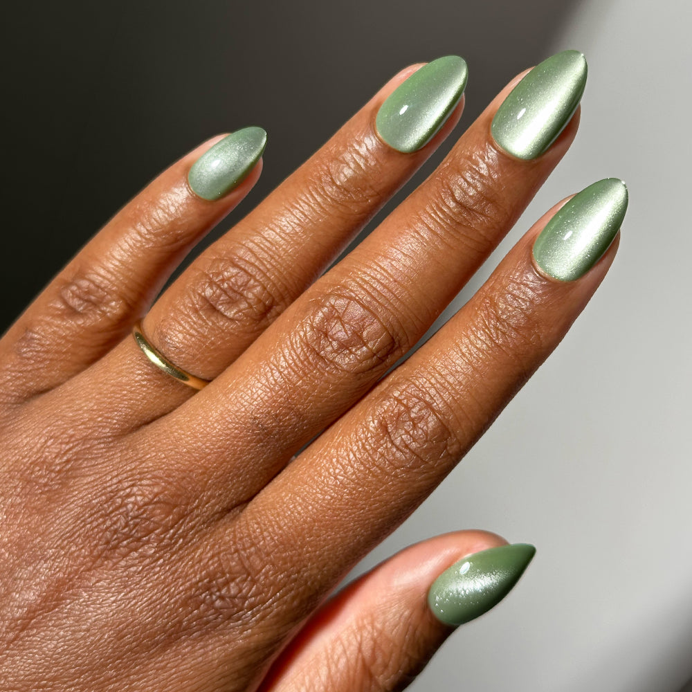 A hand with short almond nails painted in Dirty Martini, a shimmery, metallic sage green with a velvet finish.