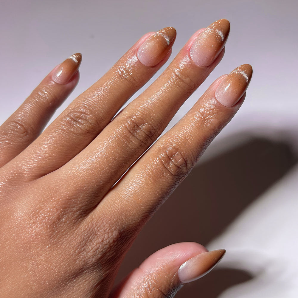 A hand with glamnetic's EARNED NOT GIVEN nude almond-shaped brown nails shows natural growth and some white powder near the cuticles. The nails feature a subtle velvet cat-eye effect, photographed against a plain background.