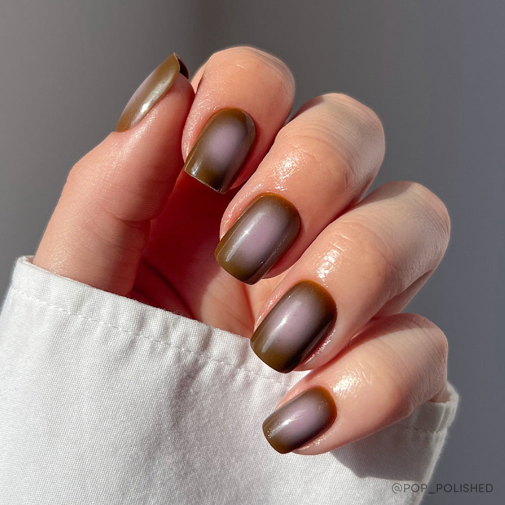 A hand with short squoval nails shows off glamnetics Eclipse, an ombre matte manicure in olive green and taupe brown. A white sleeve and sunlight highlight the chic, well-groomed look. - Product image