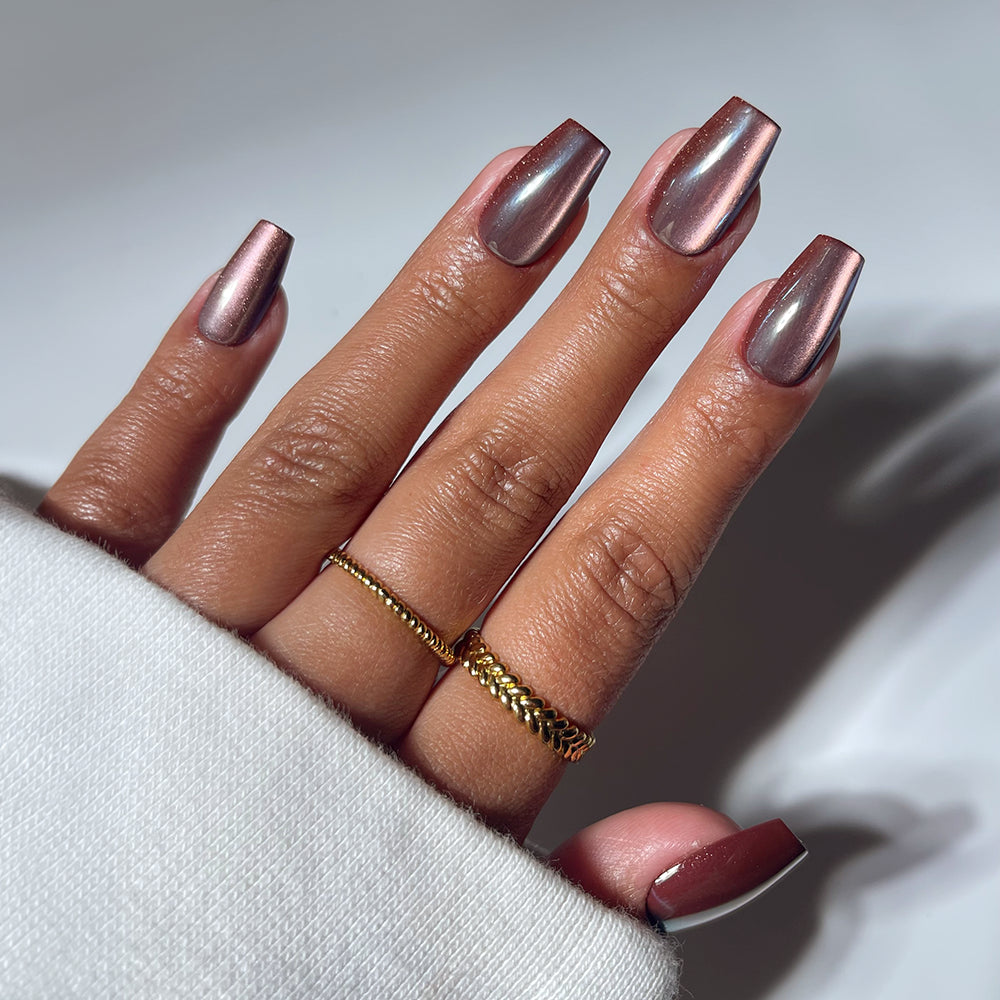 A hand with glamnetic's Espresso Glow medium coffin nails is shown, featuring metallic rose gold polish. Two gold rings adorn the fingers, and a white sleeve is visible against a softly lit plain background.