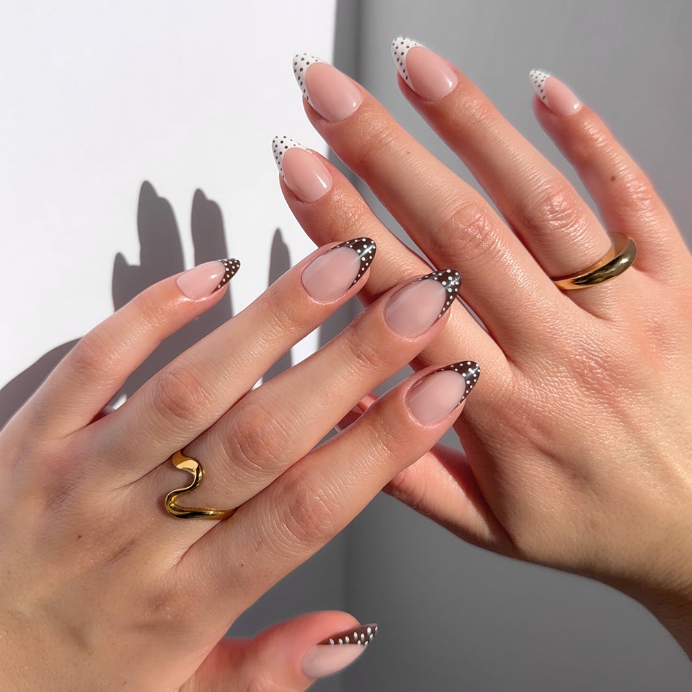 Two hands feature glamnetic's Polka In Positano almond nails with French tips—white with black dots on one hand, black with white dots on the other. Gold rings, including wavy and thick bands, accent this customizable manicure.