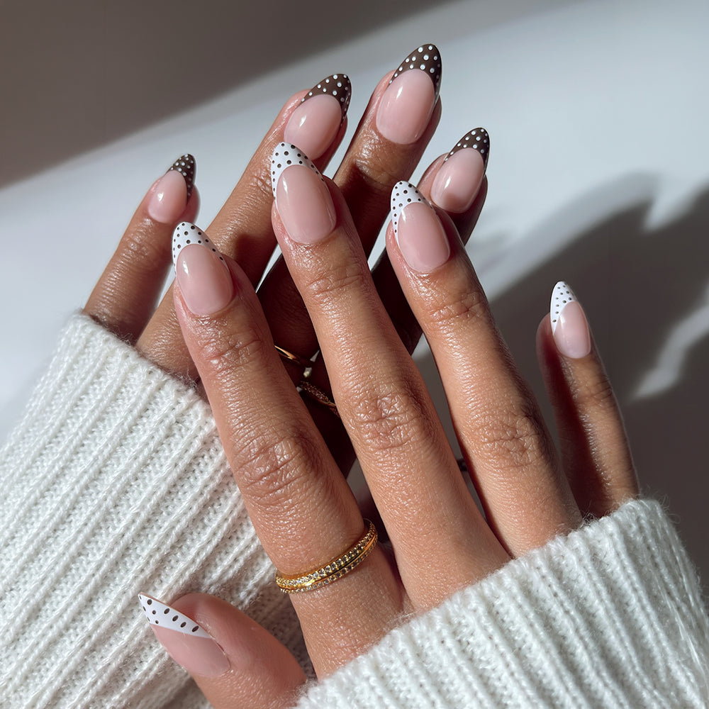 Two hands showcase glamnetic's Polka In Positano almond nails with a nude base and black and white polka dot tips. The look is styled with gold rings, a white knit sweater, and soft lighting for a chic finish. - Product image