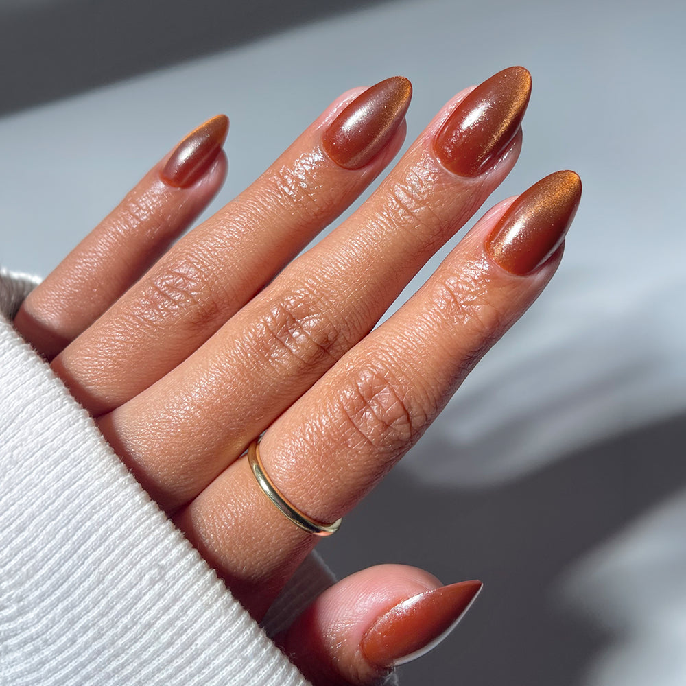 A hand wears glamnetic’s Sicilian Sun short almond press-on nails in shimmery brown, accented by a simple gold ring and a light-colored sleeve.