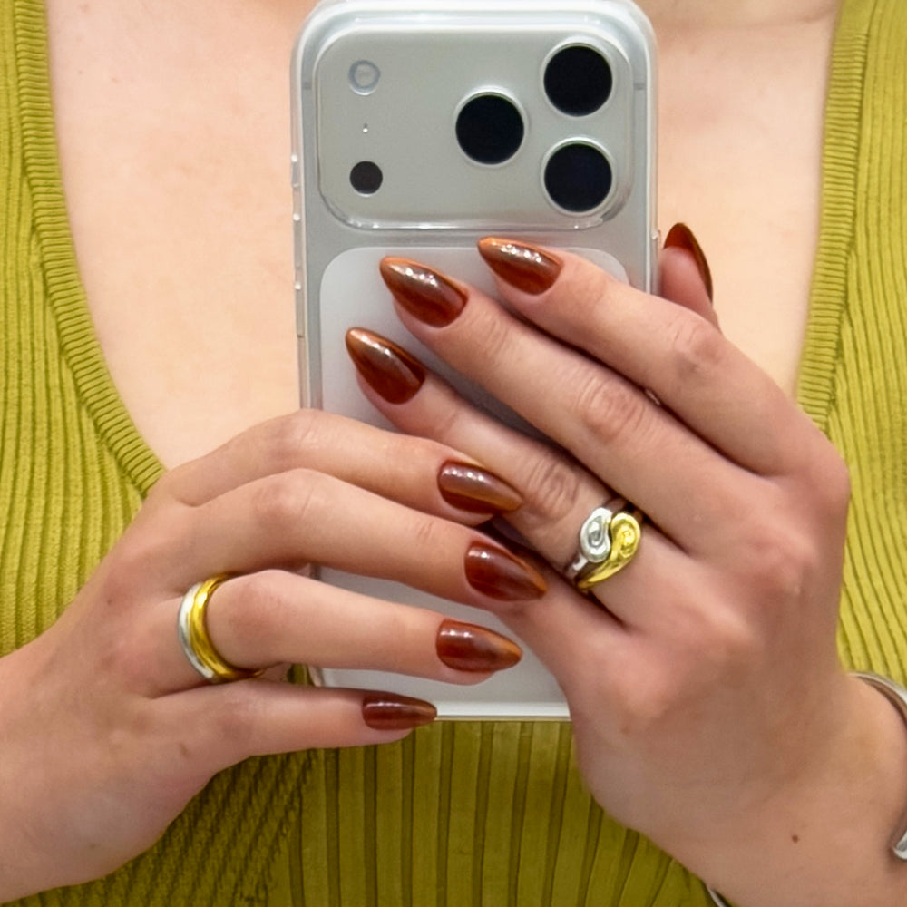 A person with long, almond-shaped brown nails—glamnetic's Sicilian Sun—holds a phone with both hands, wearing a green ribbed top and two rings: one gold and silver, the other gold with a swirl design. - Product image