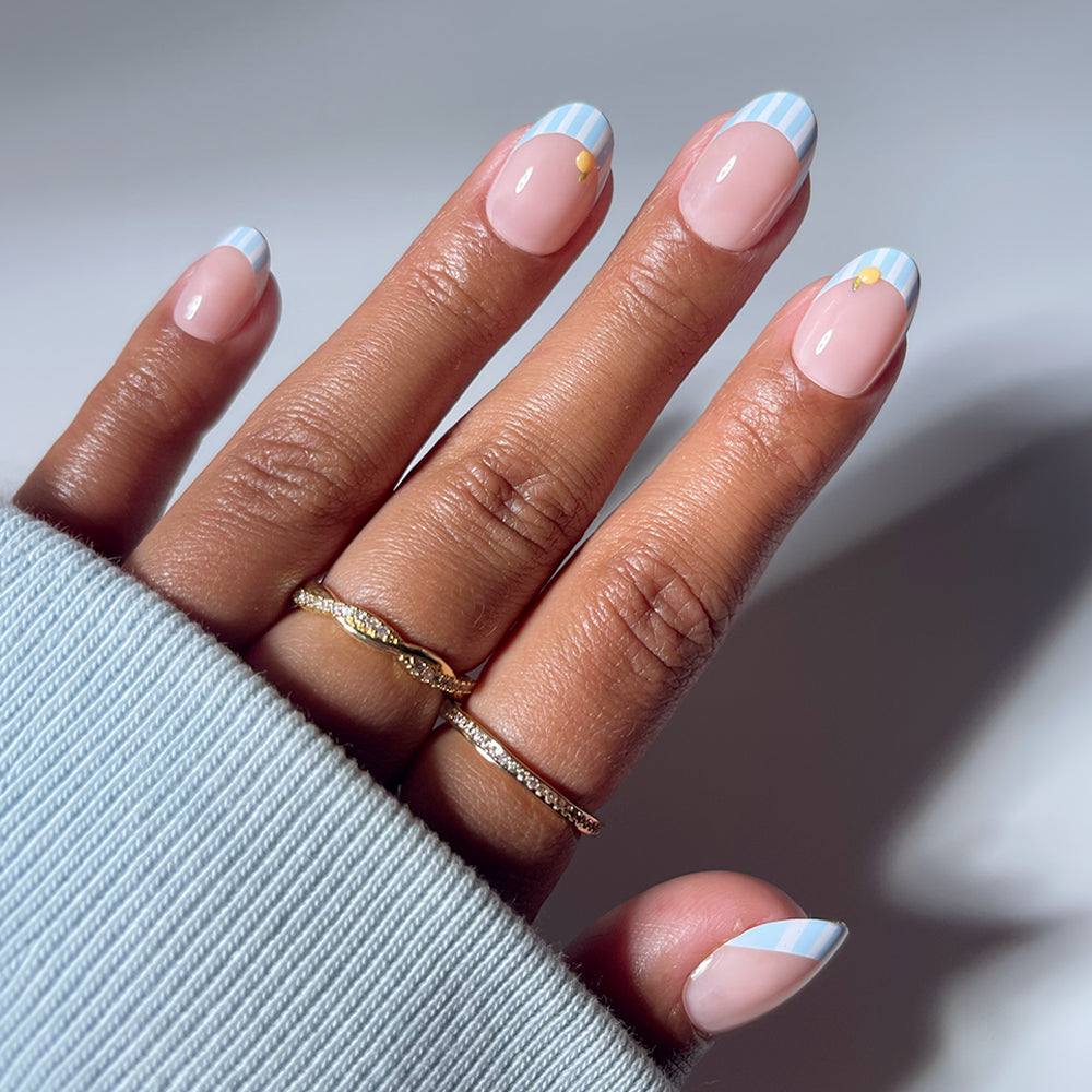 A hand showcases glamnetic's Summer Fresco: short oval nails with pastel blue and white striped French tips, each accented by a tiny yellow dot—an elegant summer look that pairs perfectly with gold rings and a light blue sweater sleeve.