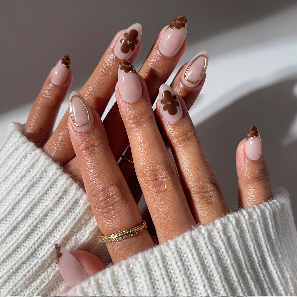 Show off salon-quality, almond-shaped nails with glamnetic’s Sunbaked—a nude polish set featuring brown abstract floral designs and gold outlines. Complete the look with a gold ring and a white knit sweater, fingers elegantly interlaced.