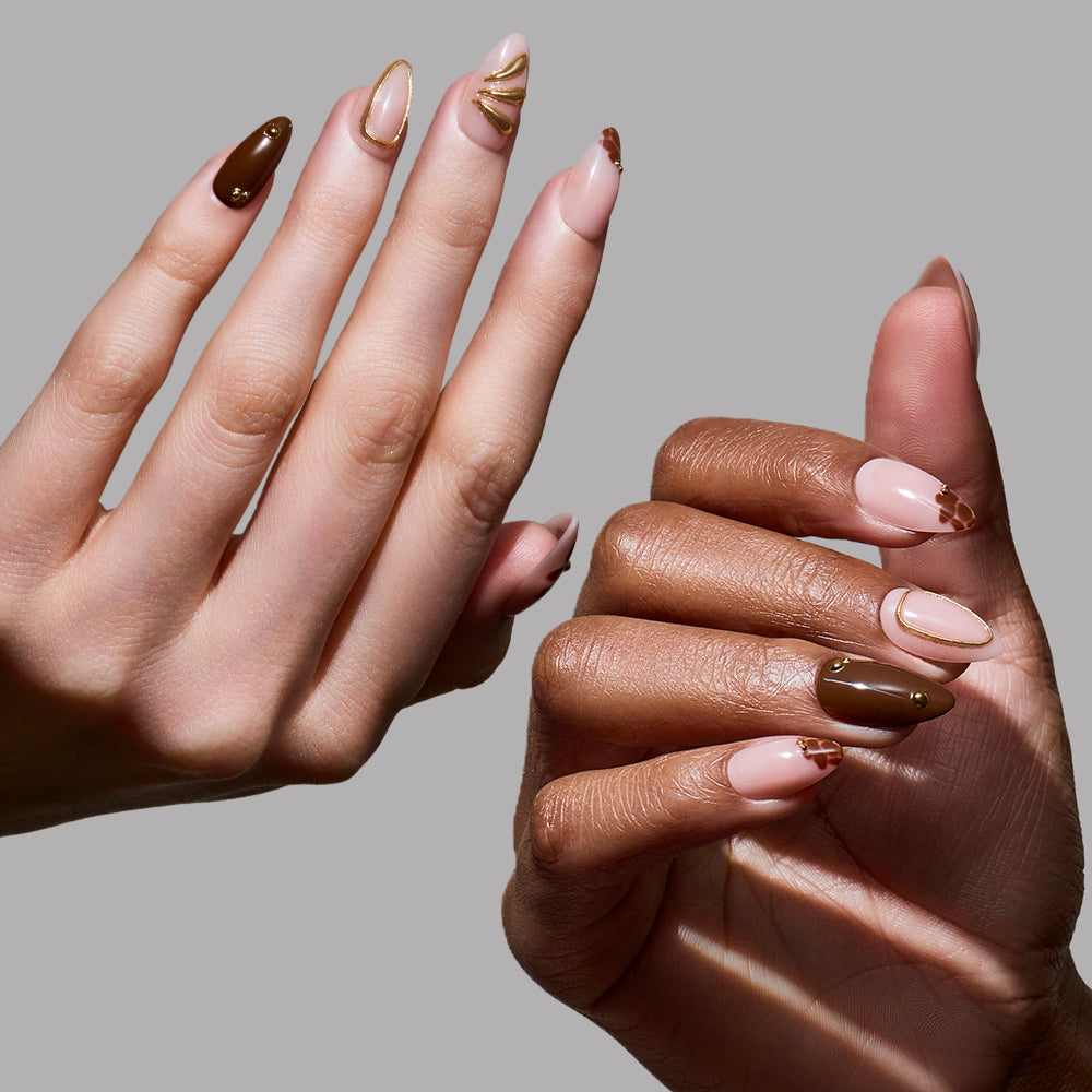 Two hands with manicured short almond nails showcase glamnetic's Sunbaked press-ons, featuring pink, brown, and gold abstract designs and swirls, set against a plain gray background. - Product image