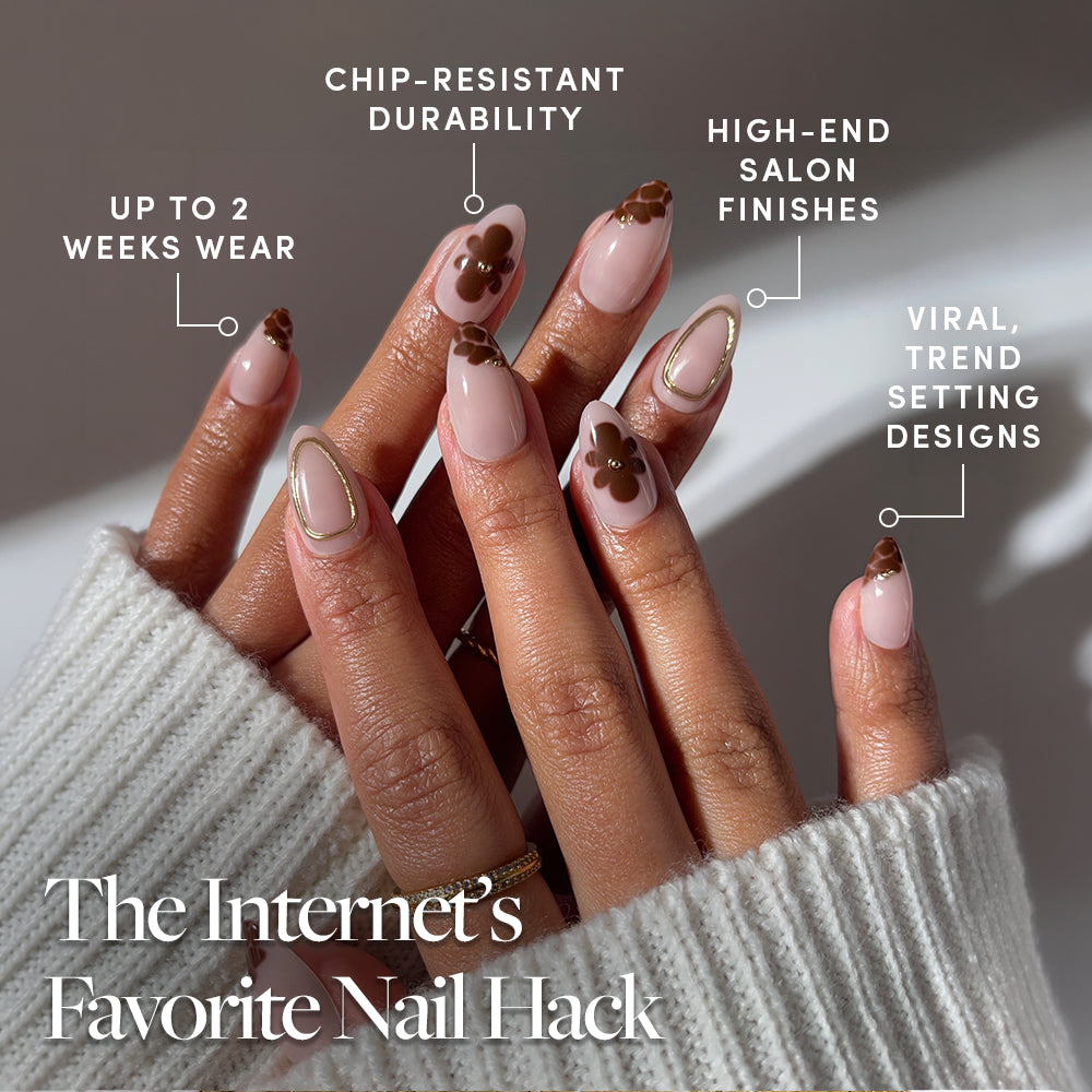 Close-up of two hands in a cozy white sweater wearing glamnetic Sunbaked nude and brown flower-themed almond press-on nails with gold accents. Features: long wear, chip-resistant, salon-quality, and trendy designs. - Product image
