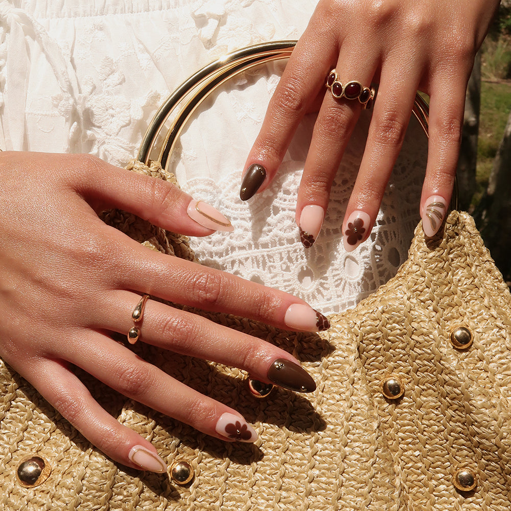 A close-up of hands wearing glamnetic's Sunbaked almond press-on nails in brown and nude floral design, accessorized with gold rings, holding a woven straw handbag with gold accents, dressed in a white textured outfit. - Product image