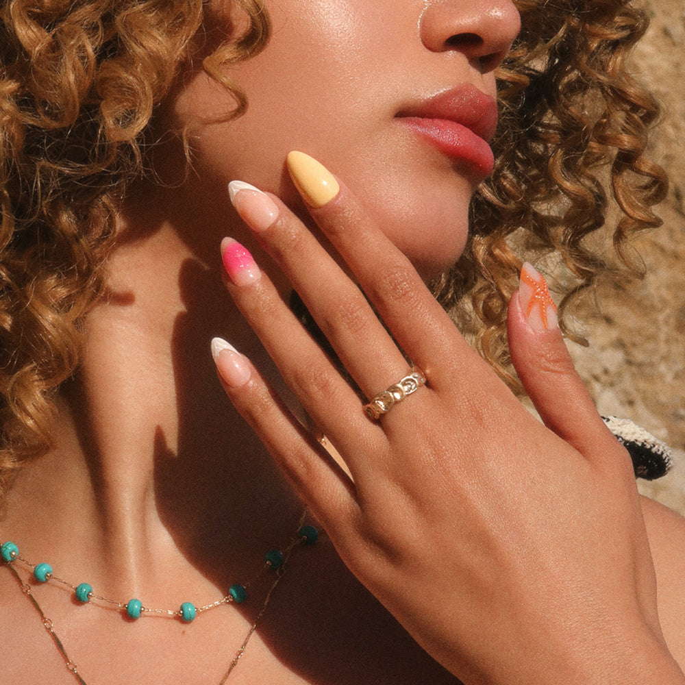 A woman with curly hair touches her face, flaunting her glamnetic Sunset Tide long, colorful manicure with a tropical design and gold rings. She wears a turquoise beaded necklace, standing in warm natural sunlight. - Product image