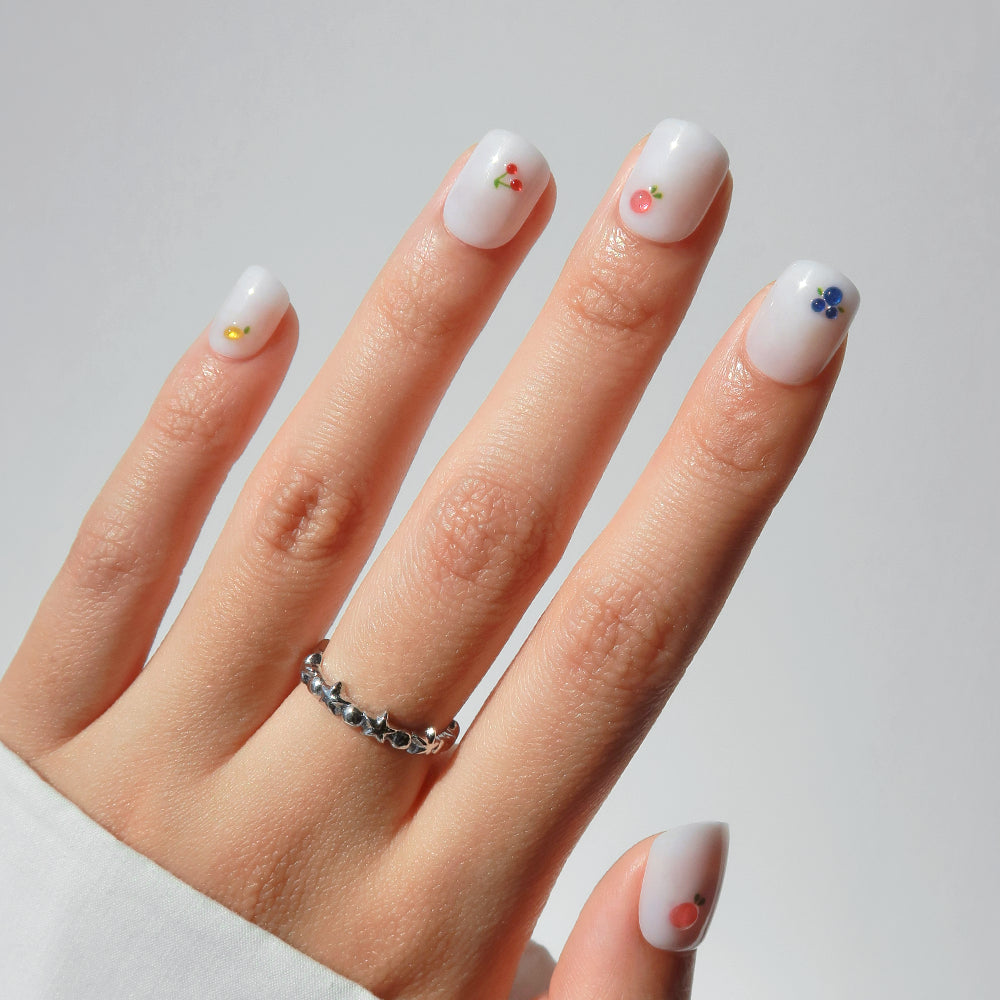 A hand models glamnetic's Sweet Picks press-on nails—short, rounded, white with tiny colorful fruit designs by the cuticle. She wears a silver ring and white long-sleeve shirt against a plain light background.