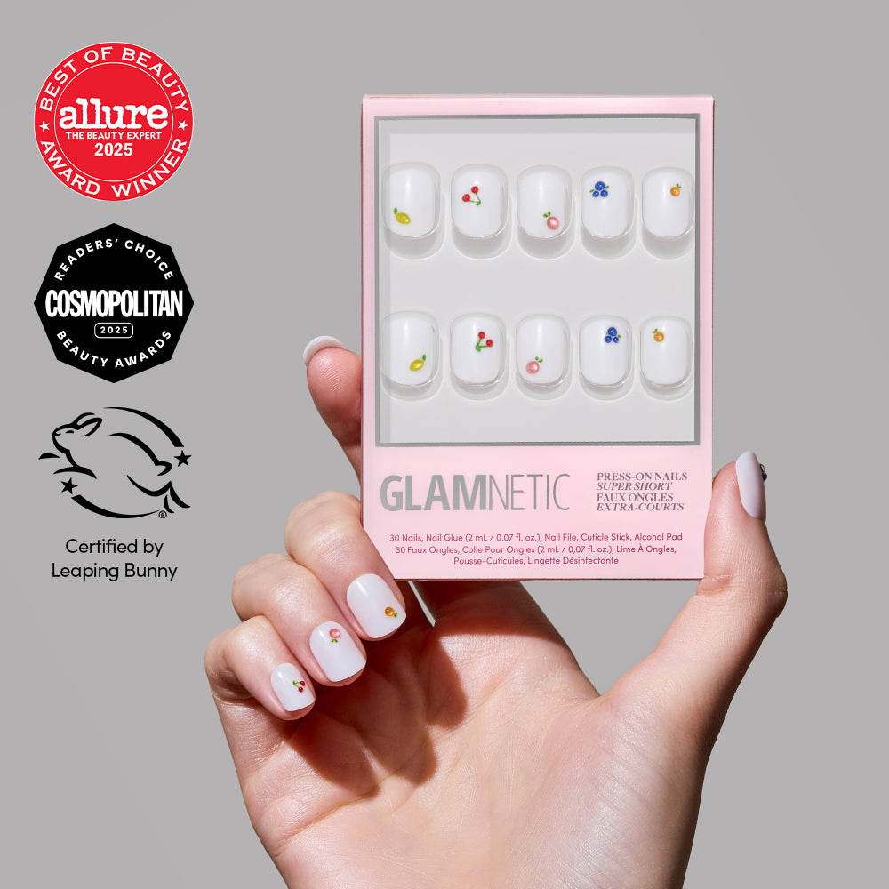 A hand with super short glamnetic Sweet Picks press-on nails, decorated with small colorful flower designs and 3D nail art, holds the product box. Award badges and a cruelty-free certification logo are shown beside the packaging.