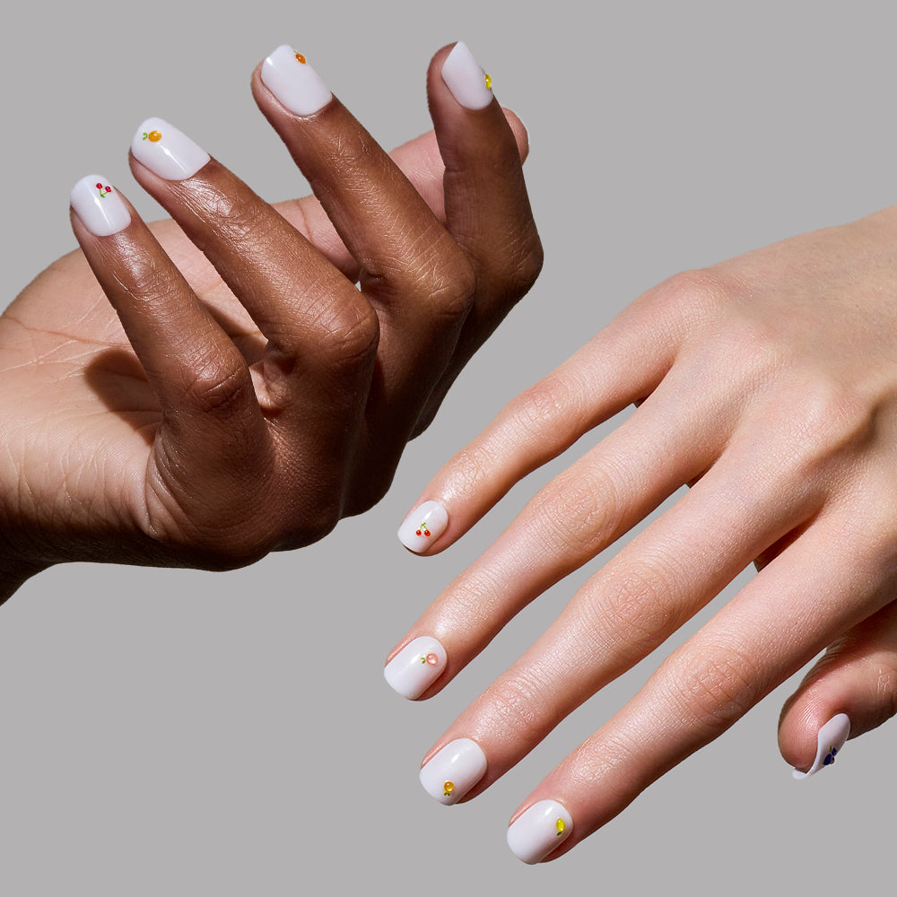 Two hands, one light and one dark-skinned, display "Sweet Picks" super short white nails by glamnetic—each accented with a single small yellow gem—against a plain gray background. - Product image