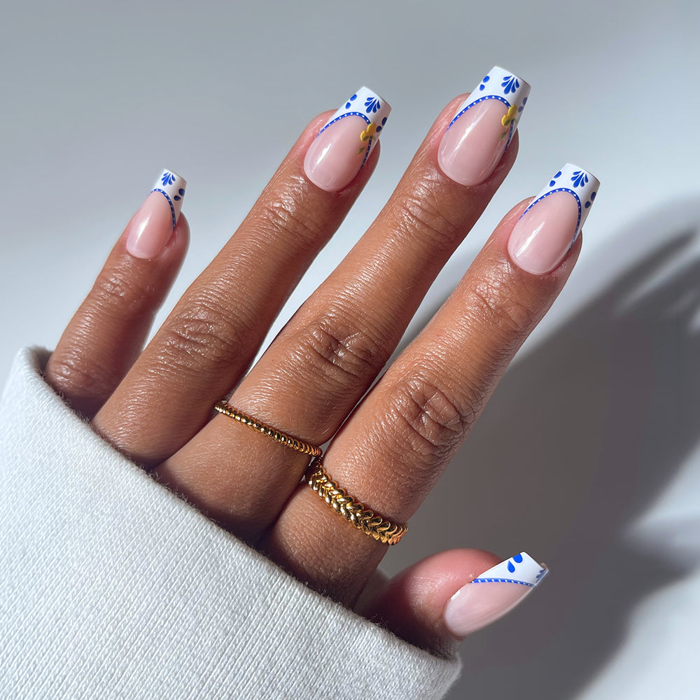 A hand with medium coffin nails in glamnetic’s Tuscan Twist style features a French manicure with white mosaic French tips, blue and yellow floral accents, two gold rings, and a white long-sleeve shirt.
