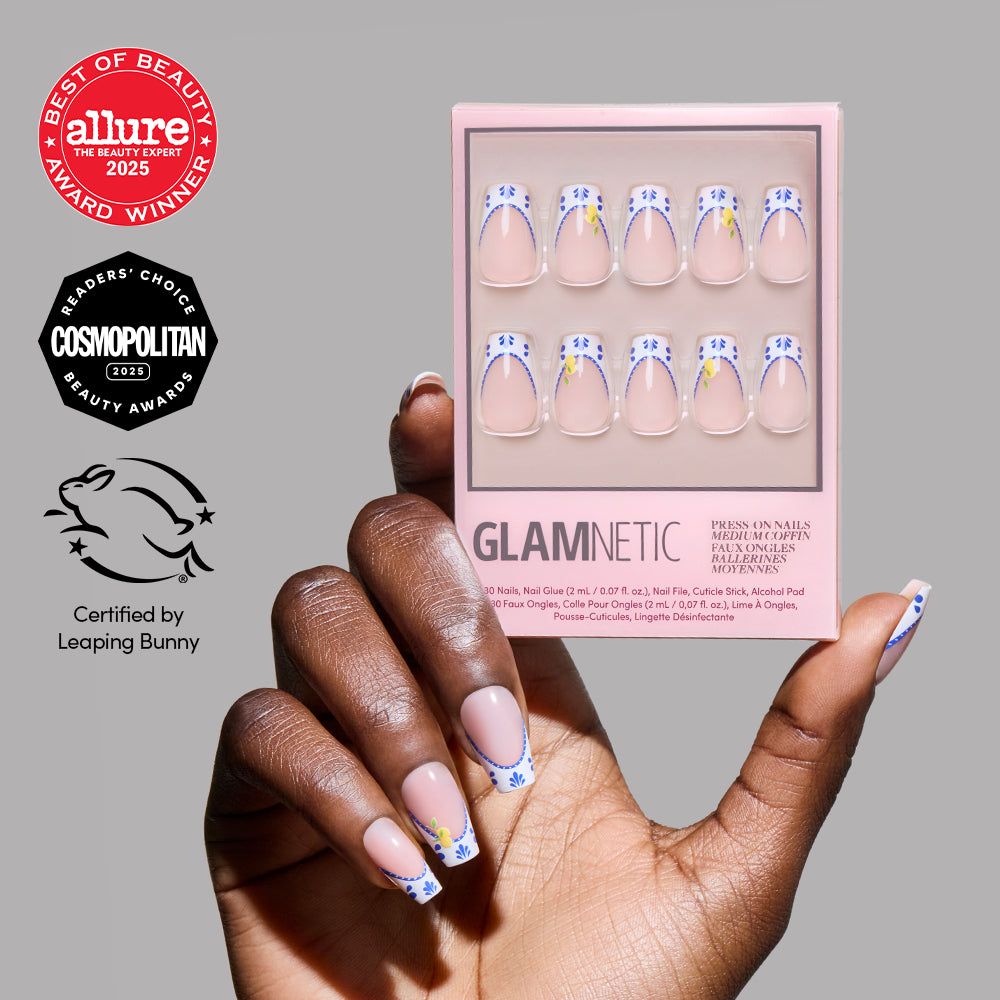 A hand with nude medium coffin press-on nails featuring blue stars and yellow accents holds a box of glamnetic Tuscan Twist nails. Allure (2025), Cosmopolitan (2023), and Leaping Bunny award badges are displayed.