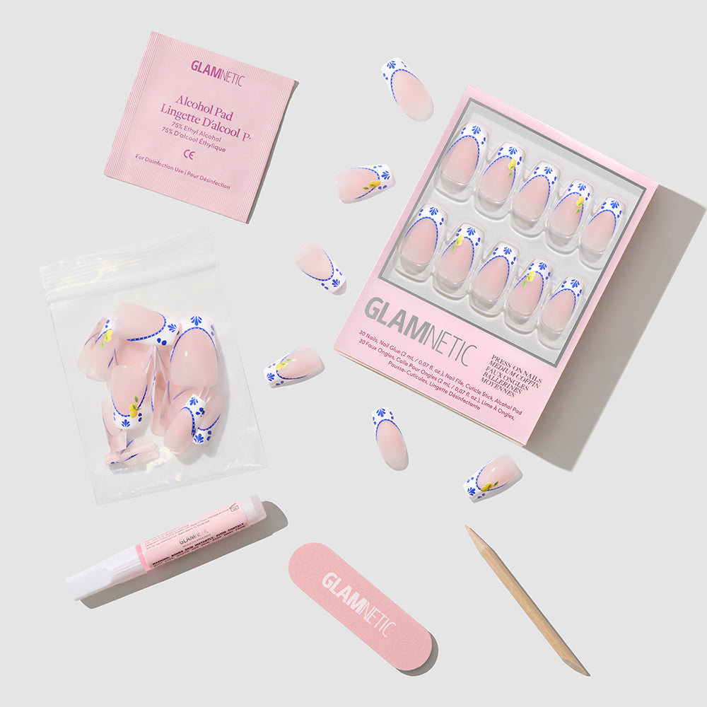 A flat lay of Glamnetic Tuscan Twist medium coffin nails kit, showing the box of pink press-ons with blue flowers, a nail file, cuticle stick, pink glue tube, alcohol pad, and extra nails in a clear bag on a light surface. - Product image