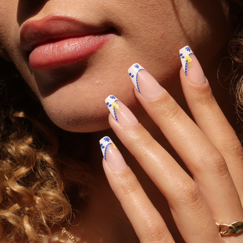 A close-up shows a woman’s lips and hand with glamnetic's Tuscan Twist medium coffin nails, featuring mosaic French tips, blue floral designs, and small yellow dots. She has curly hair and wears a gold ring. - Product image