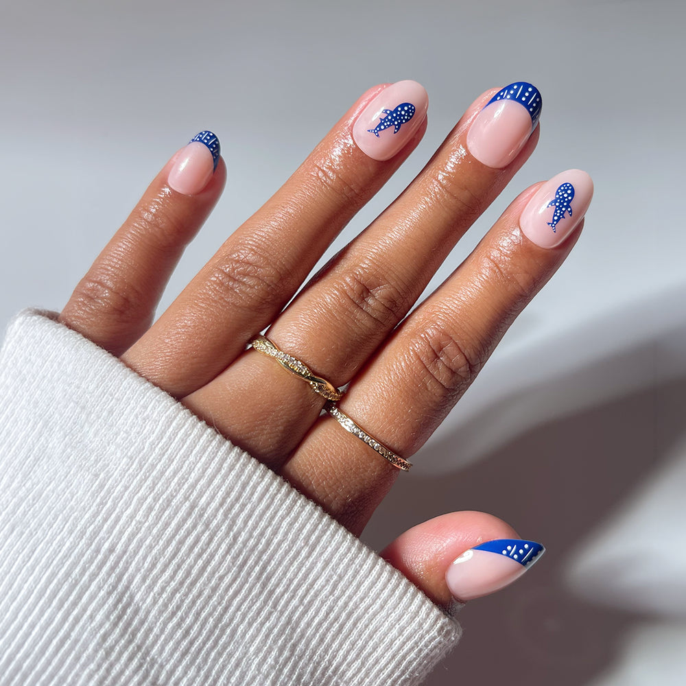 A hand with almond-shaped nails in pale pink and dark blue French tips, styled like glamnetic's Whale Waves, wears two gold rings and a cream sweater sleeve partially covers the wrist.