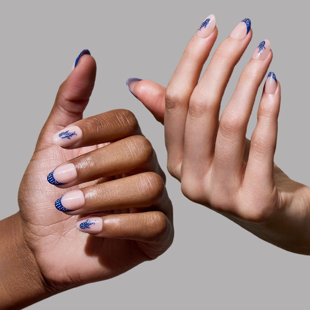 Two hands with manicured nails are shown against a plain gray background. The glamnetic Whale Waves press-on nails feature a nude base with dark blue French tips in an elegant lace-like design on a short oval shape. - Product image