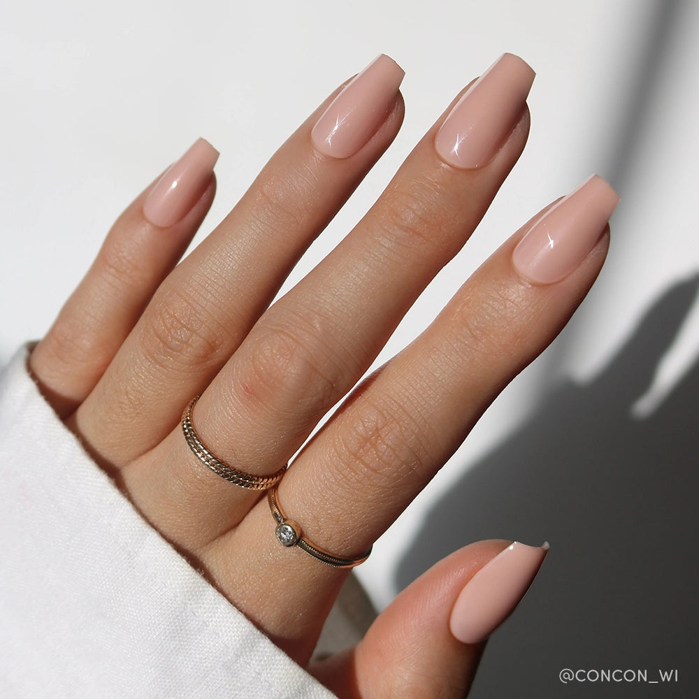 A hand with long, coffin-shaped pinky nude nails in Exposed by glamnetic, featuring a high-shine finish and two gold rings on the middle and ring fingers. A white sleeve and neutral background complete the look. - Product image
