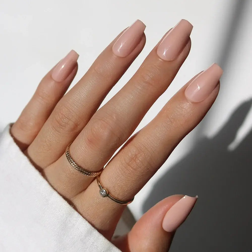 A hand with long, medium coffin-shaped glamnetic Exposed press-on nails in a pinky nude shade, wearing two gold rings—one featuring a small gemstone—is posed against a neutral background, with the person dressed in a white sleeve. - Product image