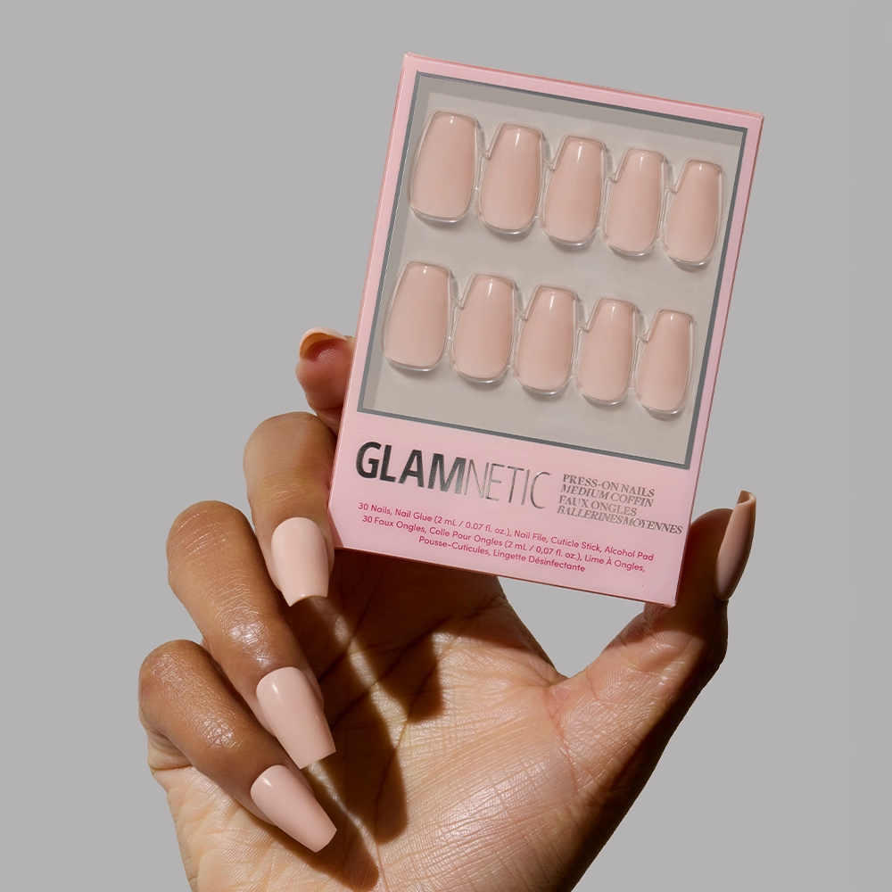 A hand with long, glossy, coffin-shaped glamnetic Exposed press-on nails in a pinky nude shade holds a matching box against a plain gray background. - Product image