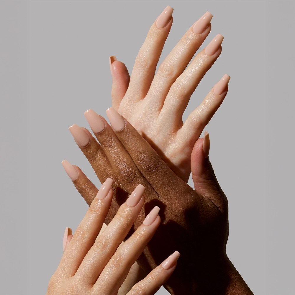 Four hands with different skin tones display long, coffin-shaped nails painted in glamnetic’s Exposed, a pinky nude polish. The overlapping pose highlights the high-shine finish against a plain gray background. - Product image