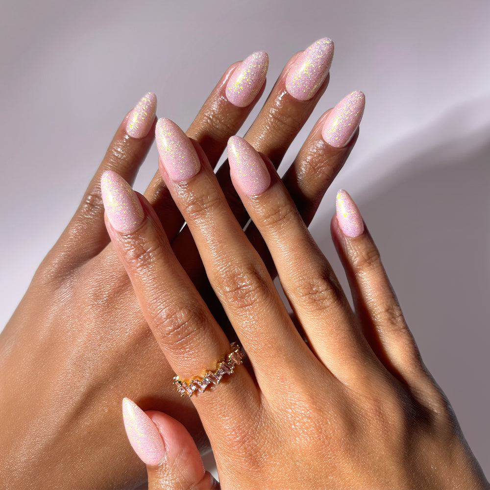 Two hands with long, almond-shaped nails wear glamnetic’s FAIRY FLEXX press-ons in a shimmery pale pink, resembling holographic shimmer nails. One hand features a gold ring with clear gemstones. Background is soft and neutral.