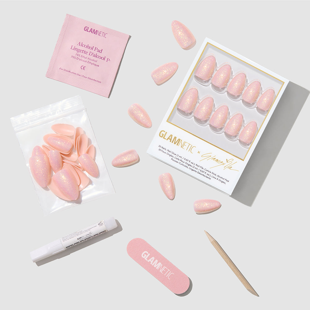 The glamnetic FAIRY FLEXX set features pink glittery short almond press-on nails with holographic shimmer, an alcohol pad, nail file, wooden cuticle stick, glue, and extra vegan cruelty-free nails arranged on a light gray surface. - Product image