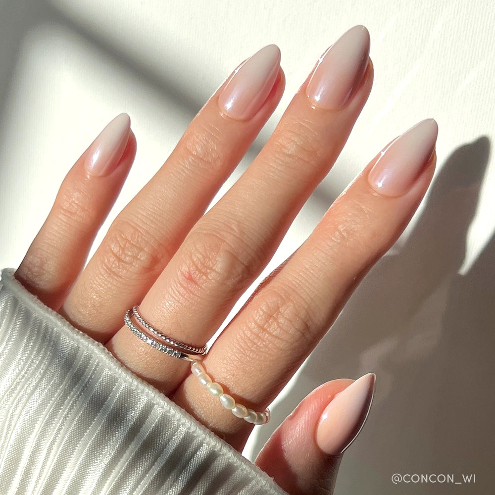 A hand with long, almond-shaped ombré nails in the shade "Fiancéd" by glamnetic, wearing two silver rings and a pearl bead ring. The person is in a white ribbed sweater, photographed under natural sunlight. - Product image
