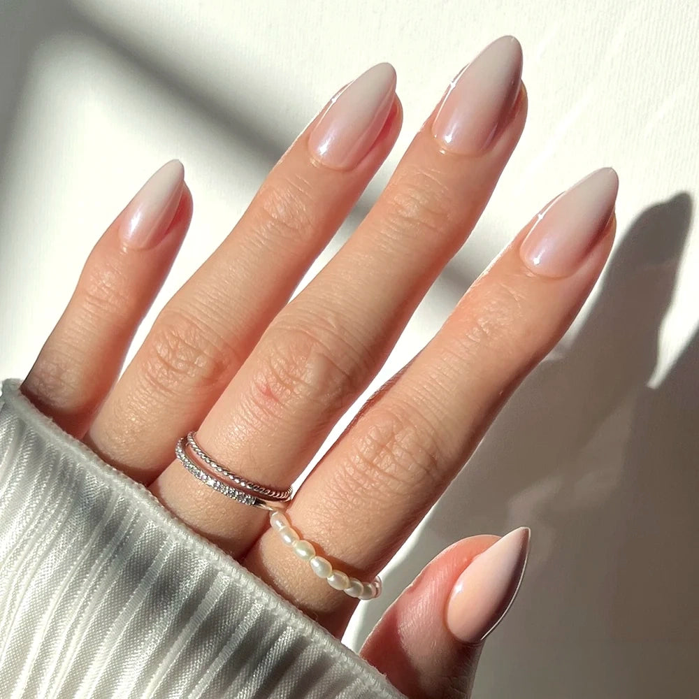 Hand with almond-shaped Fiancéd nude ombré nails featuring a delicate silver ring and a small pearl bead ring.
