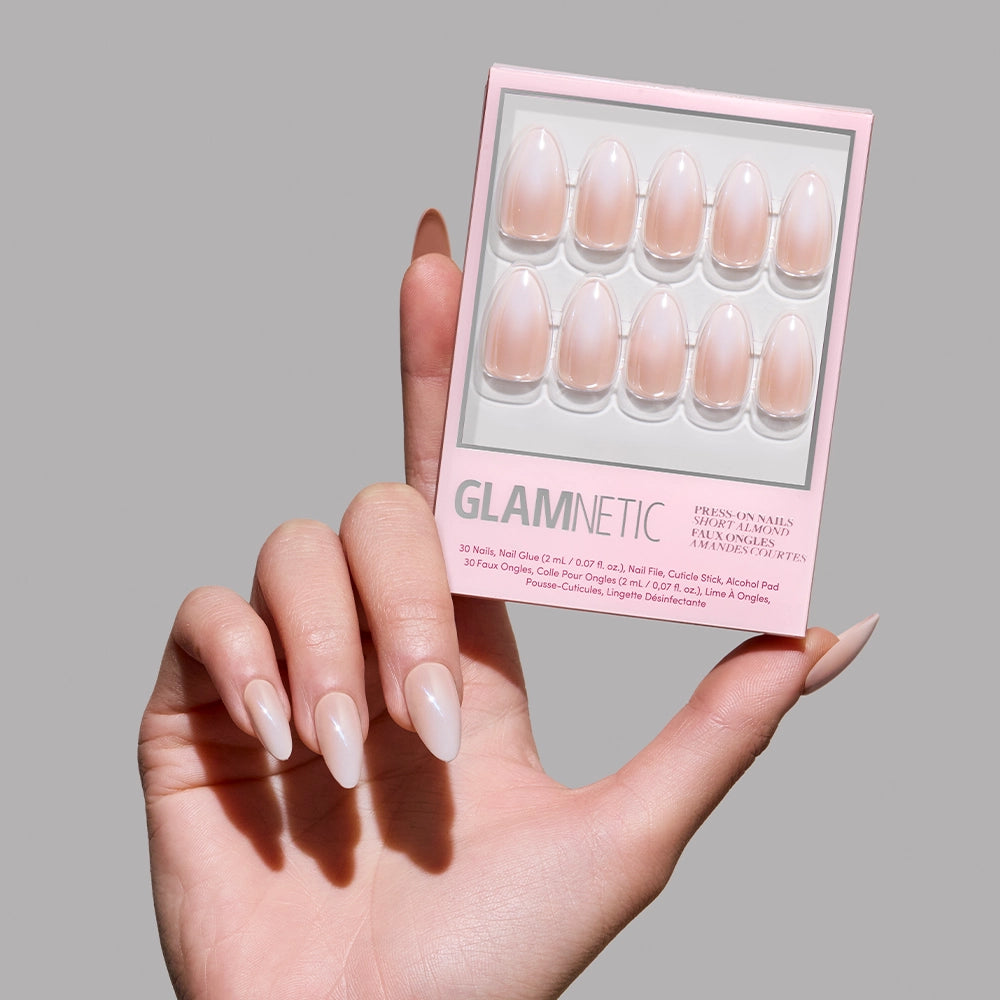 A hand with long, glossy, almond-shaped nude glamnetic Fiancéd press-on nails holds the box against a light pink background, displaying elegant French tips for a classic look. - Product image
