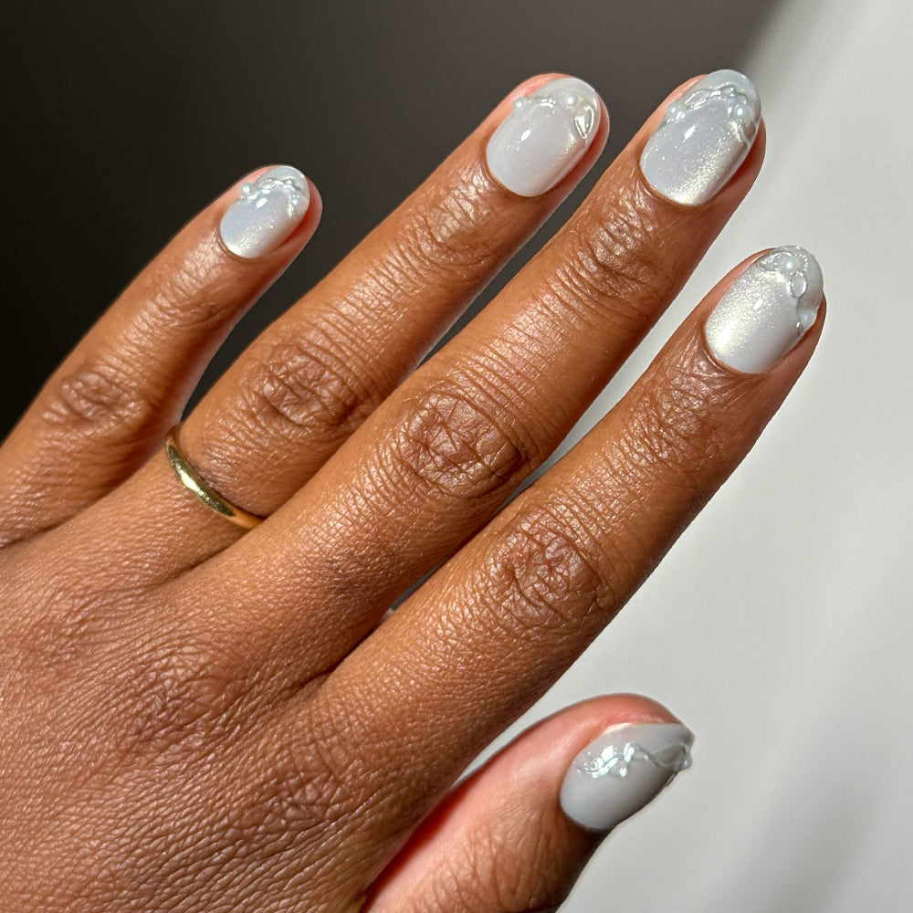 Short, rounded Fine Silver Glamnetic press-on nails painted in a shimmery, light grey polish and decorated with small white pearls and raised swirling accents.