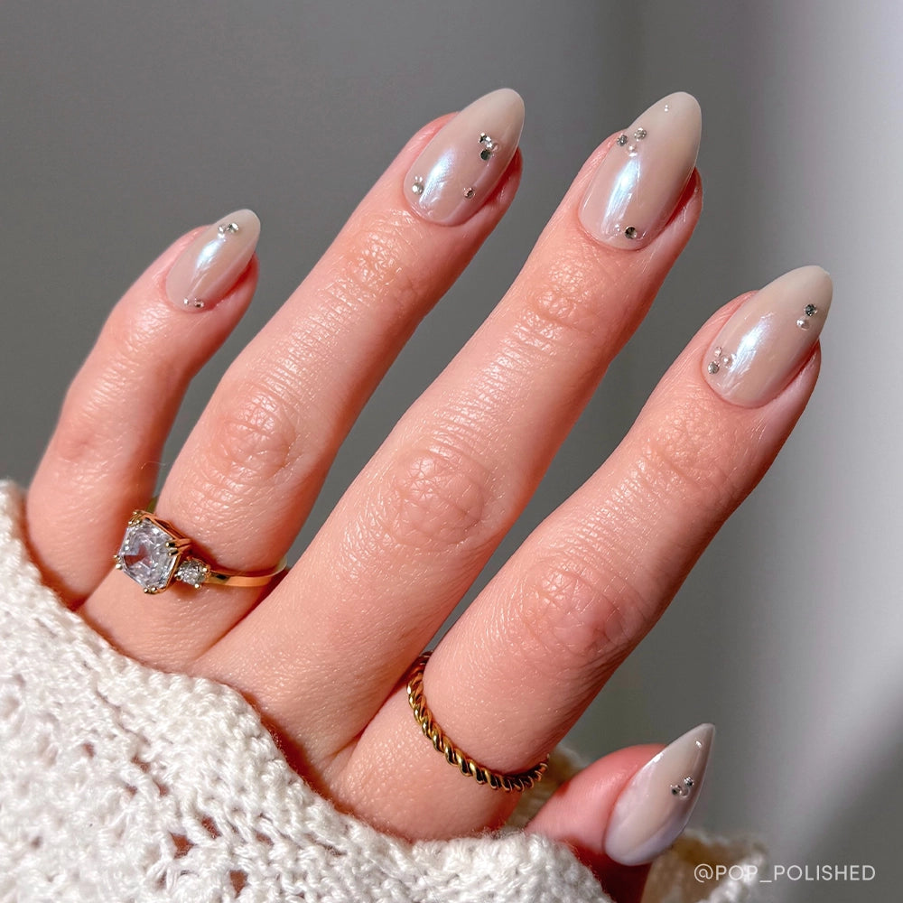 A hand showcases almond-shaped, nude glamnetic First Dance nails with small silver rhinestones, accessorized by a gold twisted ring and one with a large gemstone. The sleeve is made of white knit fabric. - Product image