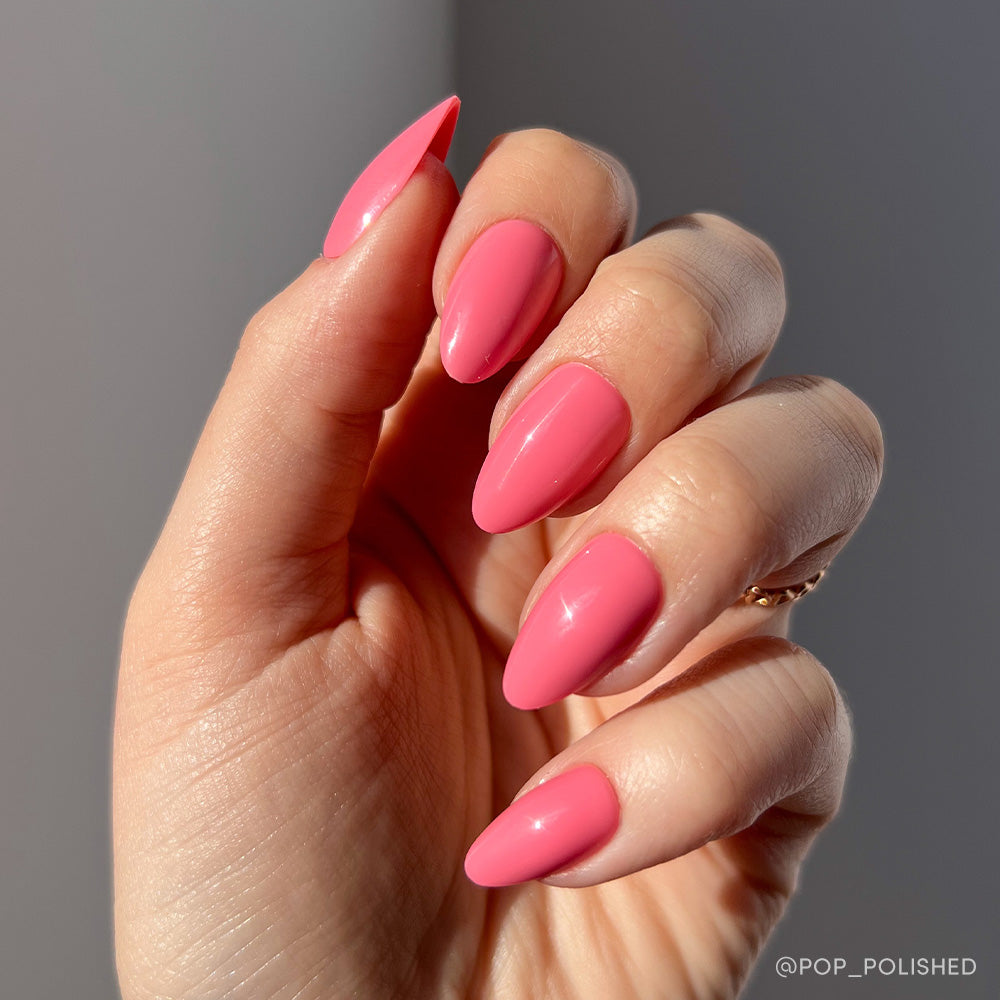 A hand with long, almond-shaped nails painted in glamnetic’s Flamingo Pink is held against a neutral background, sunlight emphasizing the glossy shine—an ideal shade for summer. - Product image