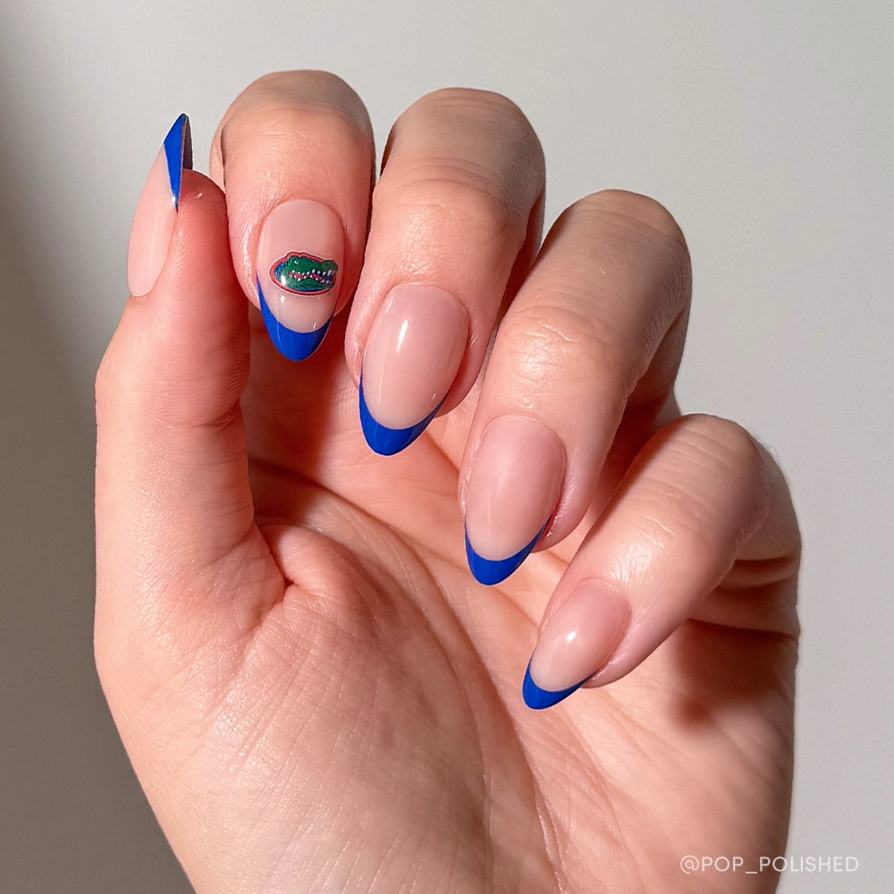 A hand sports almond-shaped nails with a nude base and bold blue French tips, just like the glamnetic Florida Gators Press-On Nail Set; the ring finger features a small, colorful, wavy cuticle design.