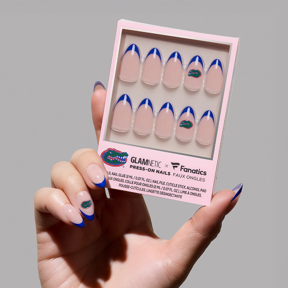 A hand displays glamnetic Florida Gators Press-On Nail Set, featuring a pink base, blue French tips, and the Gators logo on one nail. The hand also wears matching press-on nails.