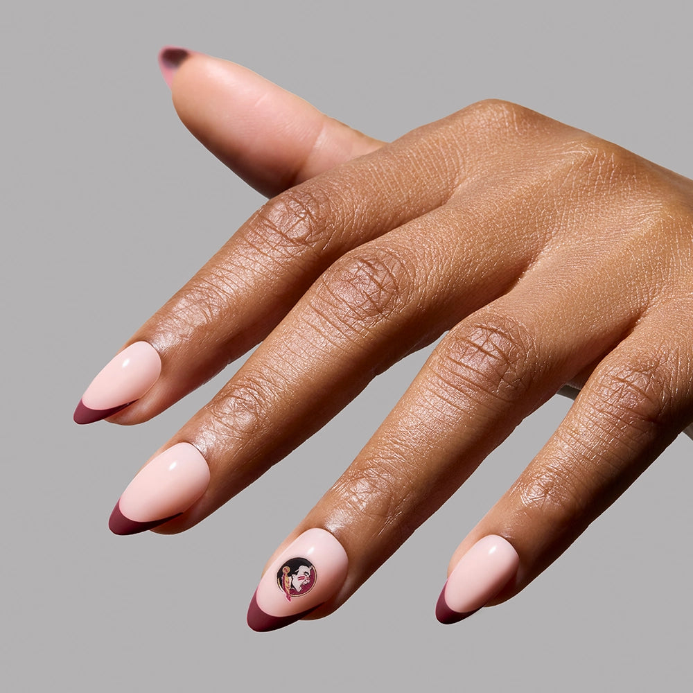 A hand wears the glamnetic Florida State Seminoles Press-On Nail Set: almond-shaped, salon-quality nails in light pink with garnet red French tips. The ring finger shows detailed nail art of a woman’s face with a bob haircut. Background is plain gray. - Product image