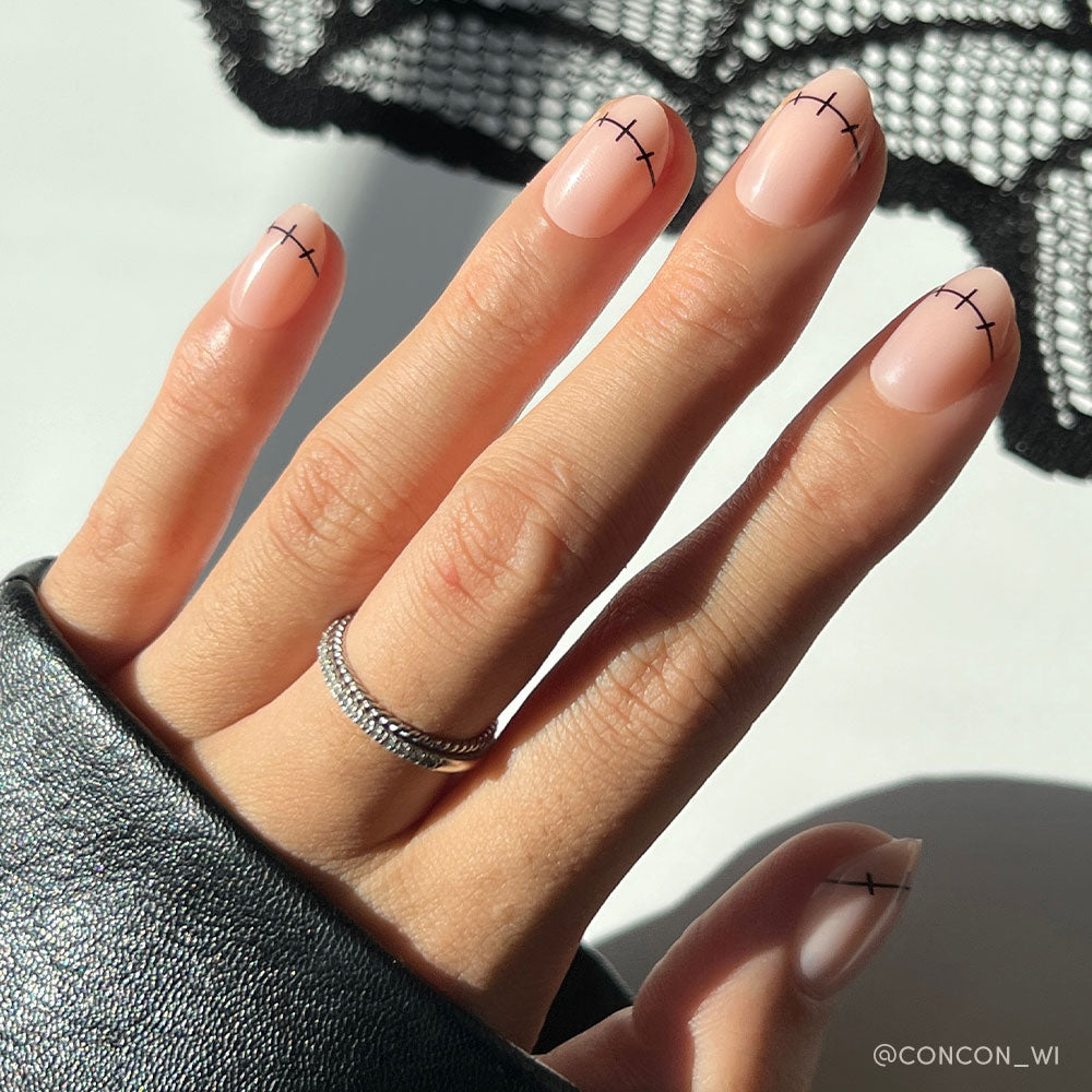 A hand with short oval nails painted in glamnetic’s Frankenbabe nude shade shows minimalist black stitch-like lines near the tips for a subtle Halloween look. A double-banded silver ring and a black leather sleeve complete the style. - Product image