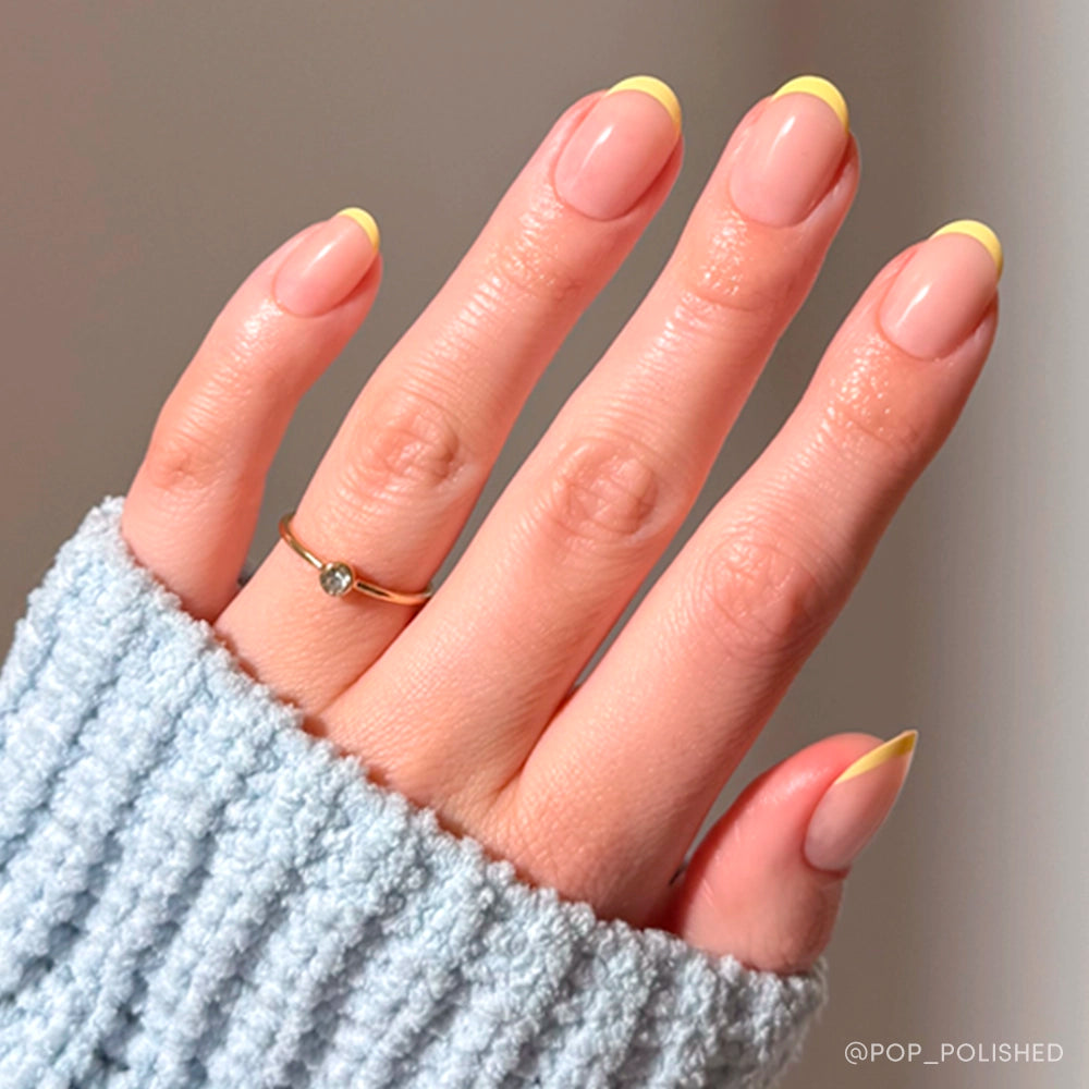 A neatly manicured hand features pastel yellow French tips from the glamnetic French Butter Press-On Nail Set, with a thin gold ring and a light blue ribbed sweater sleeve partially covering the hand. - Product image