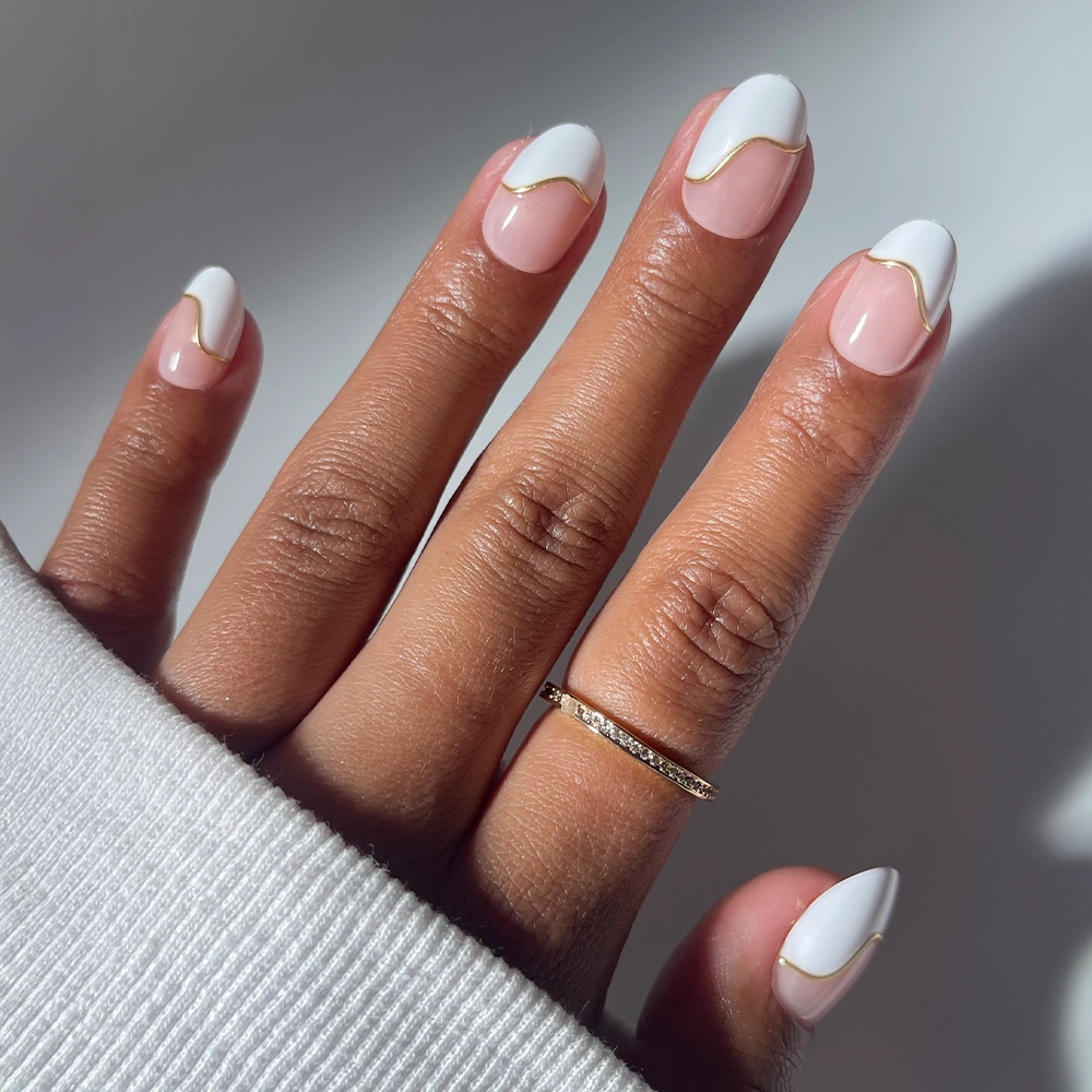 A salon-quality mani with glamnetic’s French Muse features almond-shaped nails with a nude base, white tips, and a gold wavy line. The look is paired with a thin gold ring and a white long-sleeve shirt.
