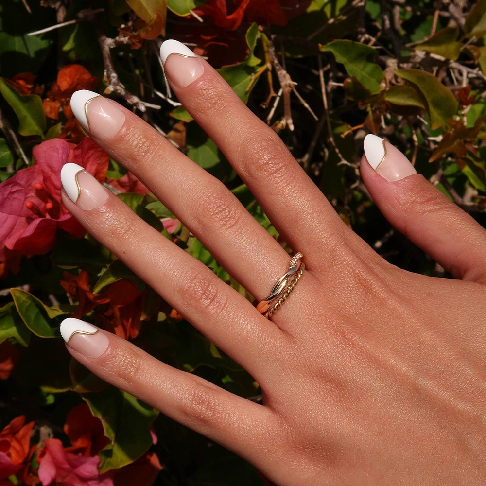 A hand with a gold ring shows off short oval French Muse nails by glamnetic, featuring white tips, against vibrant green leaves and pink flowers for a salon-quality look. - Product image