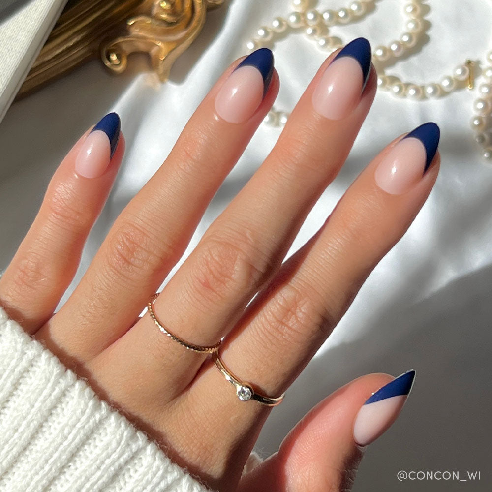 A hand with short almond nails in glamnetics French Riviera—nude base and navy French tips—wears two gold rings and a white knit sweater, with pearls and gold accents in the background. - Product image