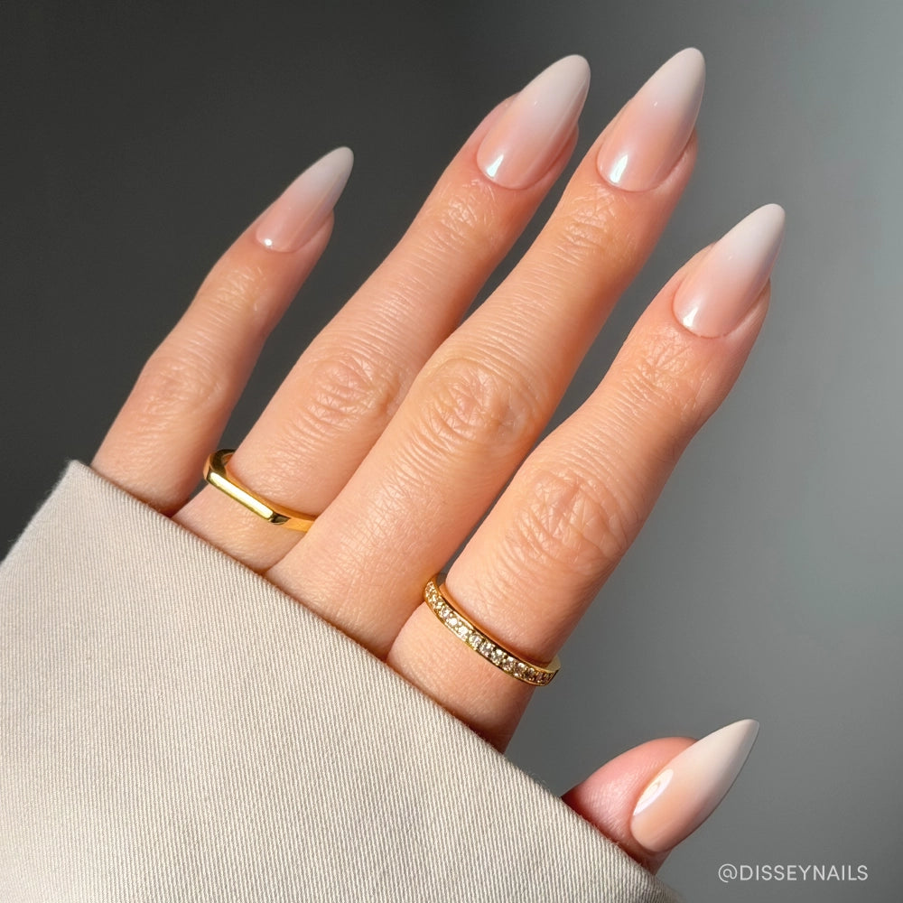 A hand with long, almond-shaped glamnetic Frosted Chai nude nails—ideal for a salon-quality look—wears two gold rings, one plain and one with small stones. The wrist is partially covered by the sleeve of a beige garment against a neutral background. - Product image