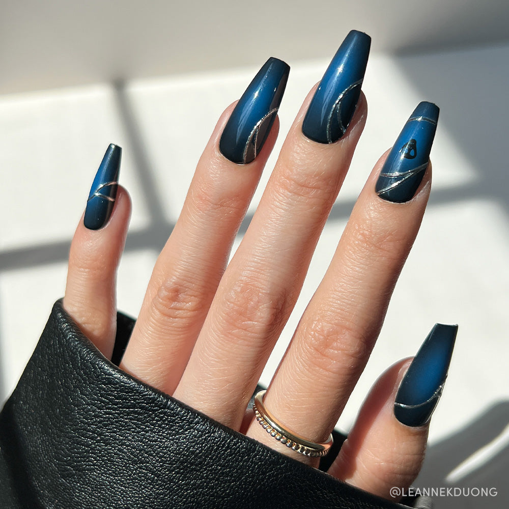 A hand with long, blue ombre coffin nails from glamnetic’s Ghost House collection features silver diagonal lines and a small heart on one nail. The look is styled with gold rings and a black leather sleeve.