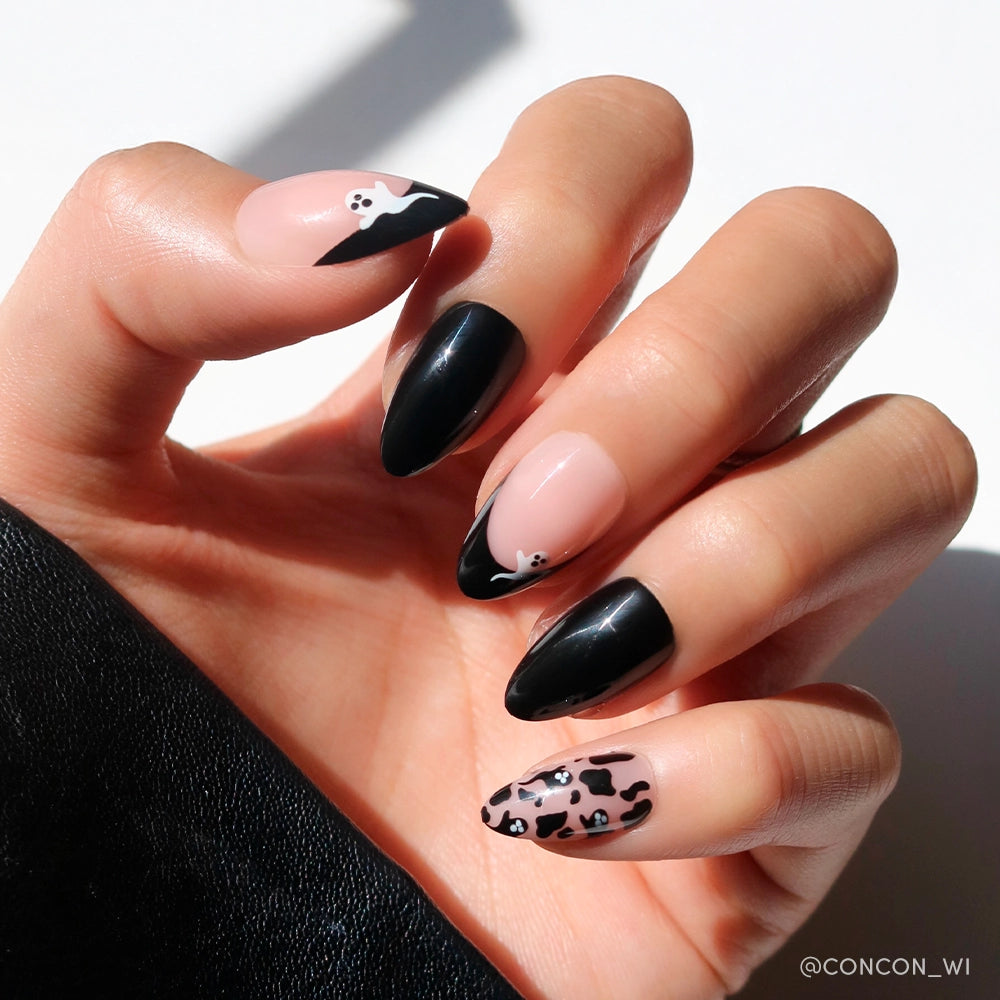 A hand displays Ghosted by glamnetic—short almond press-on nails with black, nude, and animal print designs, featuring ghost nail art like white skulls and abstract patterns. The glossy nails are set against a bright background.