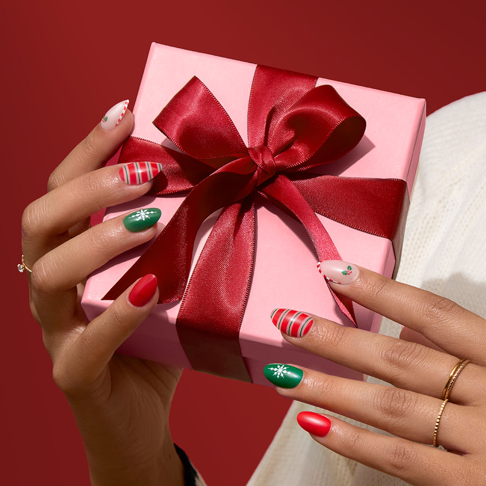 Gifting Season - Hands hold a pink gift box with a red ribbon, showcasing the Gifting Season holiday nail set on short oval nails featuring red and green stripes, snowflakes, and candy canes.