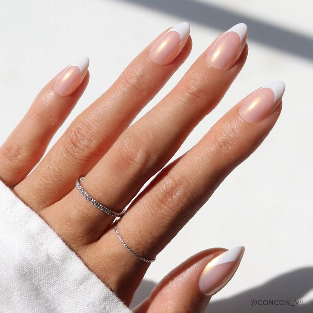 A hand showcases almond-shaped press-on nails with a luminous champagne pearl overlay and crisp white French tips. The nails have an iridescent glow in natural sunlight, and the hand is accessorized with two dainty silver rings.