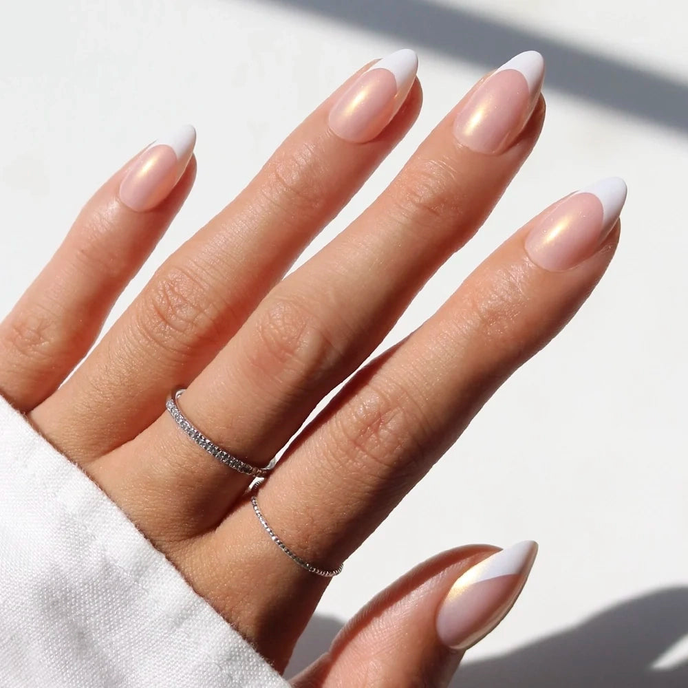 A hand with long almond-shaped GIVE ’EM GRACE press-on nails by glamnetic features a pearly pink base and elegant white French tips. The person wears two thin silver rings, and the cuff of a white sleeve is visible.