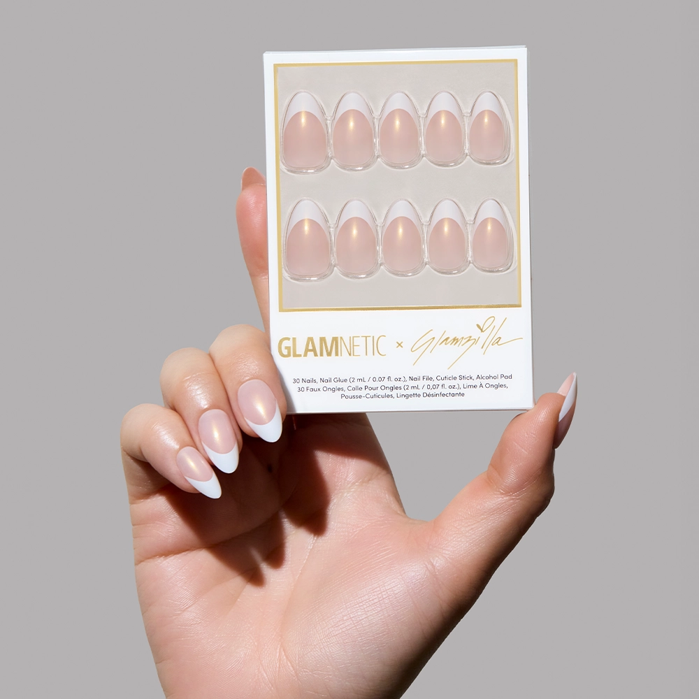 A hand holding the “Give ‘Em Grace” nail set in its packaging. The box features 30 nails in a soft blush tone with a pearlescent sheen and bold French white tips, arranged neatly in a clear tray with Glamnetic and Glamzilla branding.